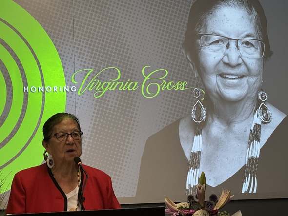 Virginia Cross speaks at a podium with her portrait and the words “Honoring Virginia Cross” displayed behind her.
