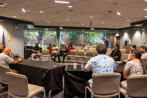 People seated in a meeting room with large windows, engaged in a community discussion.