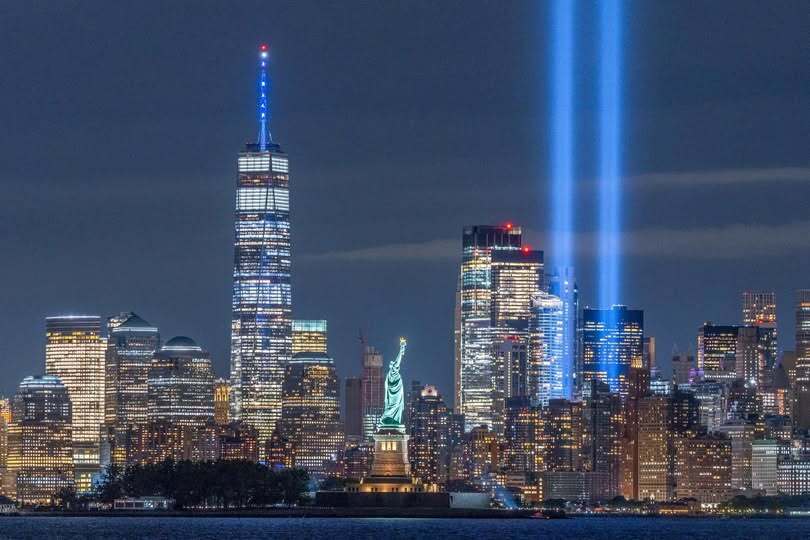 New York City skyline at night with Tribute in Light beams and Statue of Liberty in foreground.