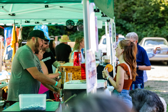 Vendor pours a drink as a smiling woman chats with him at a festival booth.