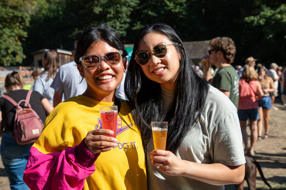 Two women smiling with sunglasses, holding drinks at an outdoor event.