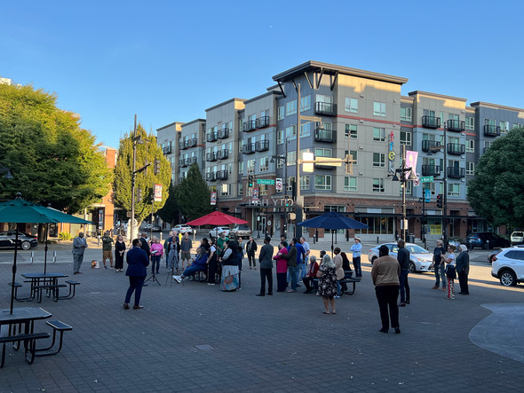 People gather in a downtown plaza near picnic tables, listening to a speaker under a clear evening sky.