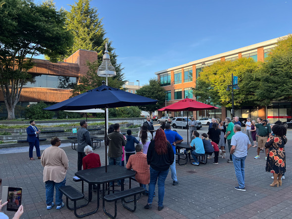 Crowd facing a speaker in a blue suit during an outdoor event, with red and blue umbrellas nearby.