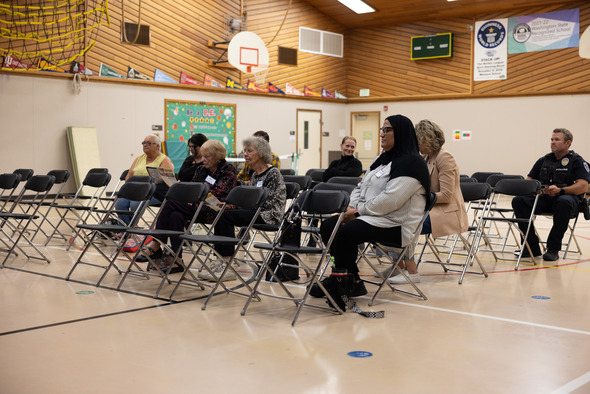 Small group of attendees seated in folding chairs listening during the community meeting in a school gym.
