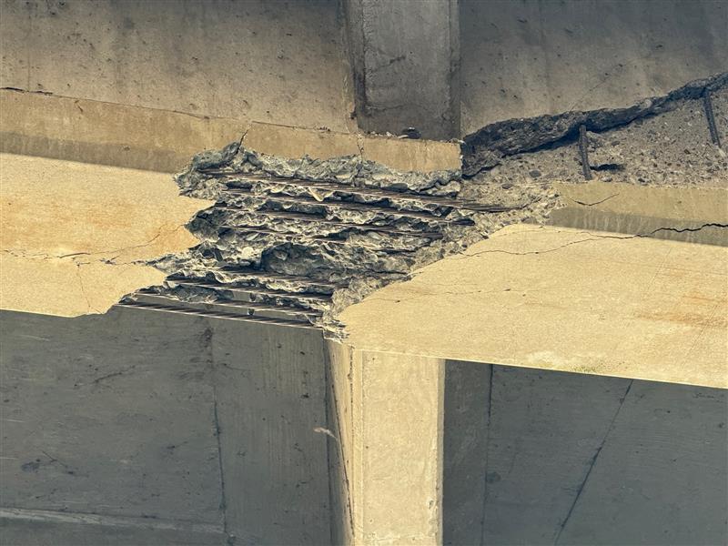 Close-up of damaged concrete bridge support showing exposed rebar and crumbling structure.