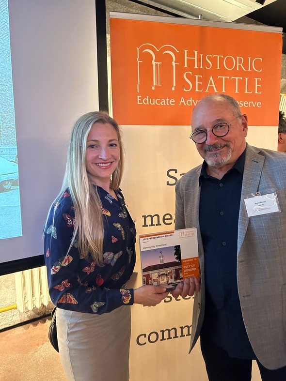 Two people standing together smiling, holding City of Auburn preservation award in front of Historic Seattle banner.