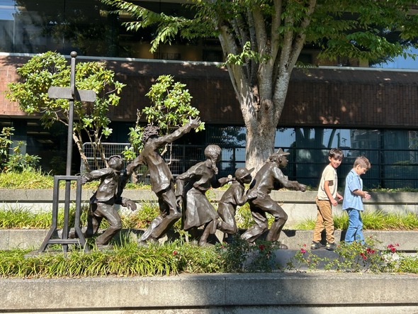 Bronze statue of children playing in a line near City Hall with two young boys walking past on a sunny day.