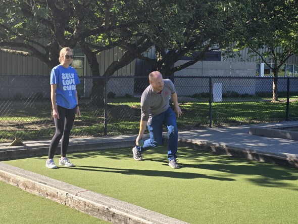 Man in gray shirt tosses bocce ball while woman in blue “Self Love Club” shirt watches.
