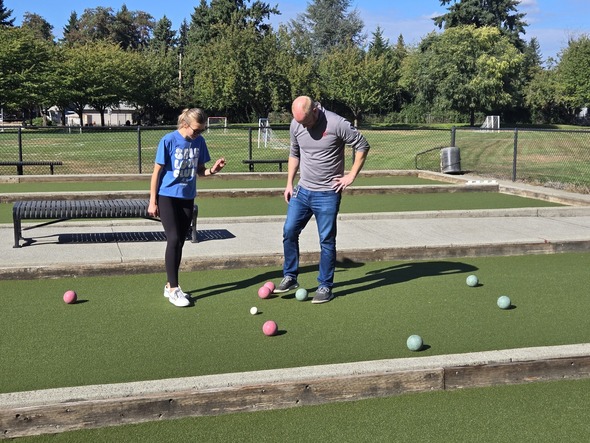 Two people stand over bocce balls on the court, checking which ball is closest.