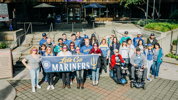 A group of people standing in a plaza holding a sign that says Let's Go Mariners dressed in Mariners gear 