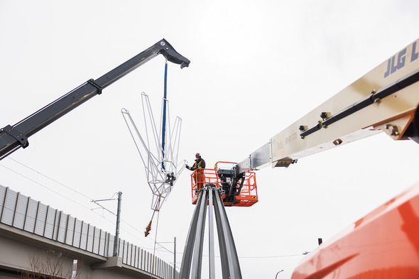 Workers use cranes to lift and install a large metal sculpture near an overpass.