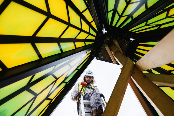 Worker in safety gear installs a metal-and-glass art structure with glowing yellow panels.
