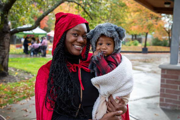 Woman in red hooded cape holds baby dressed as a wolf during a Halloween event outdoors.