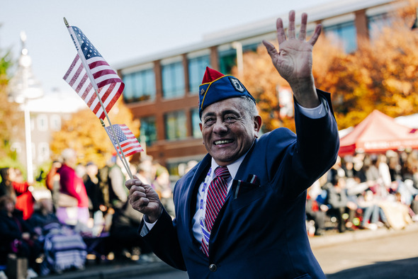 Veteran in a blue suit smiling and waving while holding small American flags in a parade.
