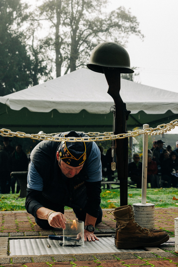 Veteran kneeling to light a memorial flame beside a battlefield cross display during a ceremony.