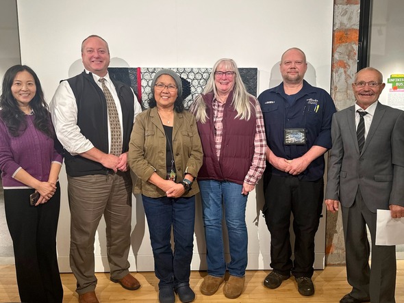 Group photo of six adults standing side by side in an art gallery, smiling at the camera.