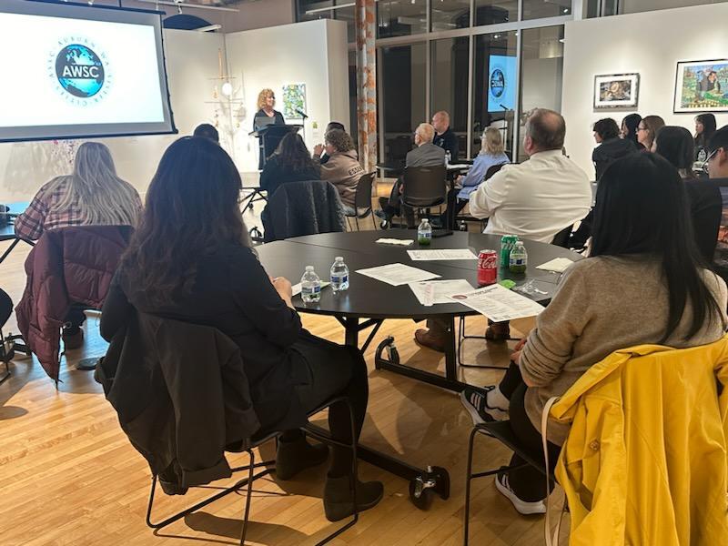 Audience seated at round tables listens to a speaker at a podium during a Sister Cities meeting in an art gallery.