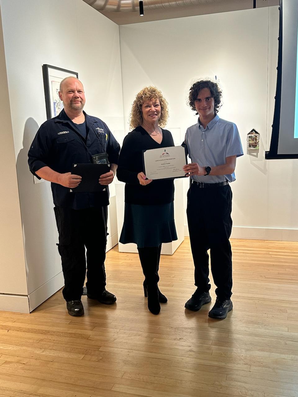 Mayor presents a certificate to a young award recipient, standing alongside another presenter in an art gallery.