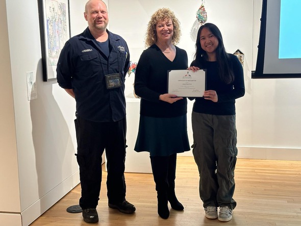 Mayor presents a certificate to a young woman, standing with another presenter in an art gallery.