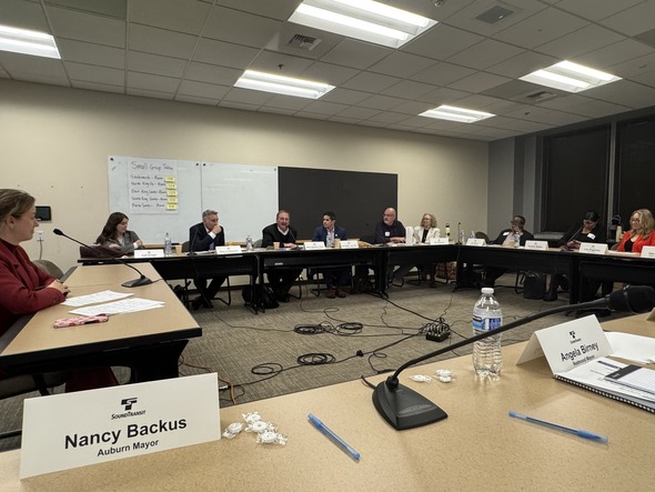 Elected officials sit in a large meeting room for a Sound Transit discussion, with nameplates and microphones arranged on the tables.