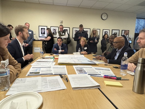 Local and regional leaders gather around a table reviewing documents during a briefing, with others standing and observing in the background.