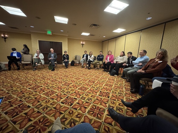 A large group sits in a circle in a conference room, participating in a discussion during a breakout session.