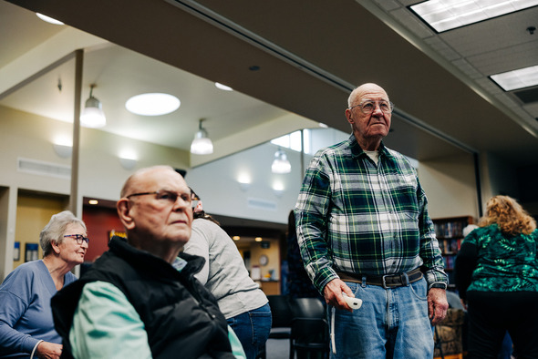 A man standing holding a Wii mote controller while another man sits in a chair to his right