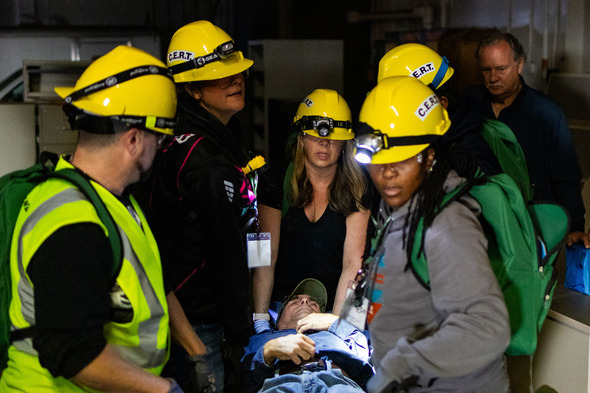 CERT volunteers in yellow helmets carry a man on a stretcher during an emergency response drill.