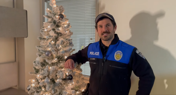 Auburn police officer stands beside a lit, flocked Christmas tree decorated with silver ornaments.