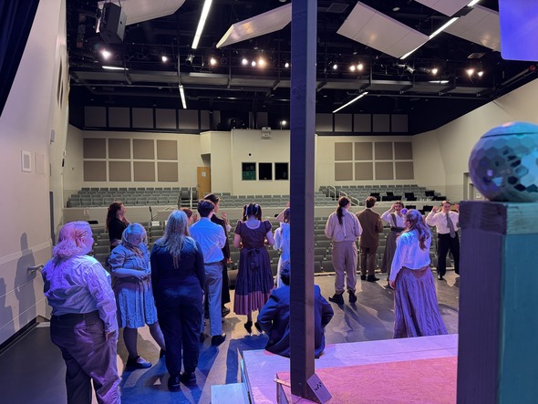 Cast members in period costumes gather onstage during rehearsal in an empty theater lit with overhead stage lights.