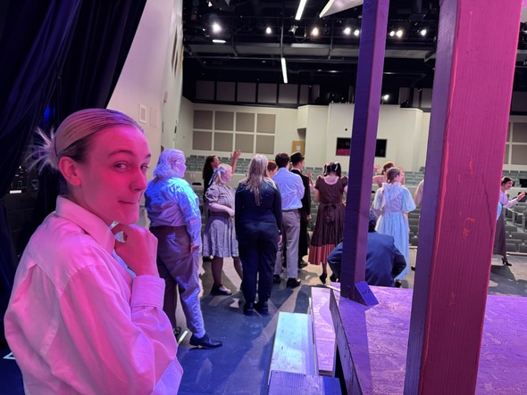 Student in stage makeup and costume stands backstage as a group of cast members gathers onstage under colorful theater lights.