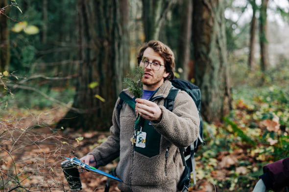 Outdoor educator holds a small evergreen branch while speaking to a group in a wooded park.