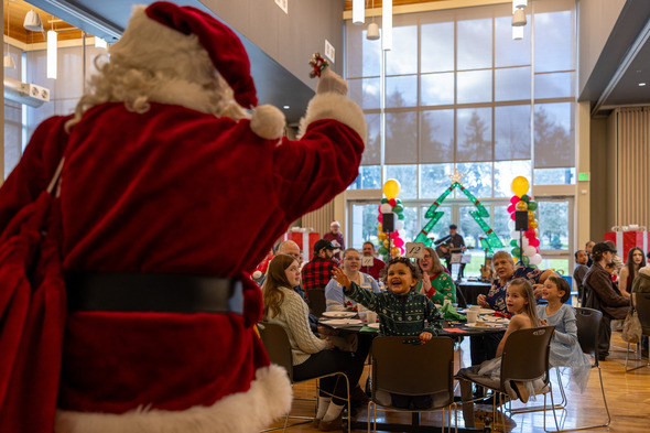 Santa waves to excited children and families during a festive breakfast event in a decorated community hall.