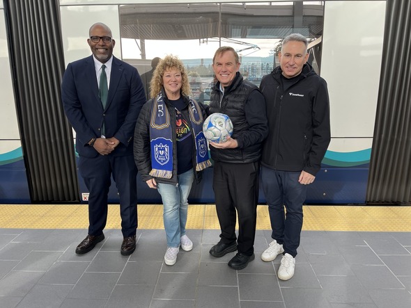 Four officials stand on a Link light rail platform posing for a photo, one holding a soccer ball.
