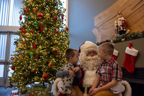 Two young boys sit with Santa beside a large decorated Christmas tree and fireplace stockings.
