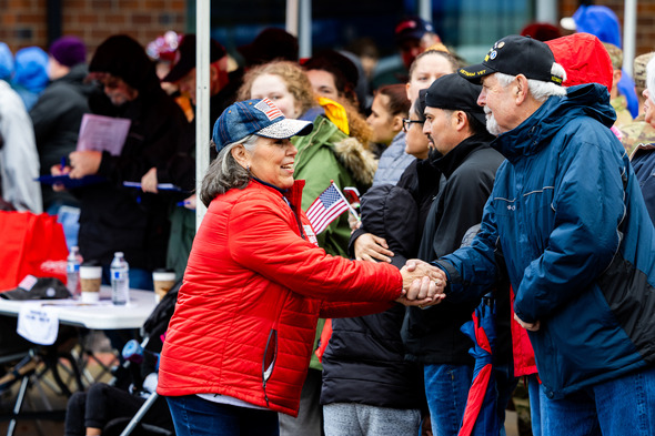 Woman in a red jacket shaking hands with a man in a crowd during a patriotic outdoor event, holding a small American flag.