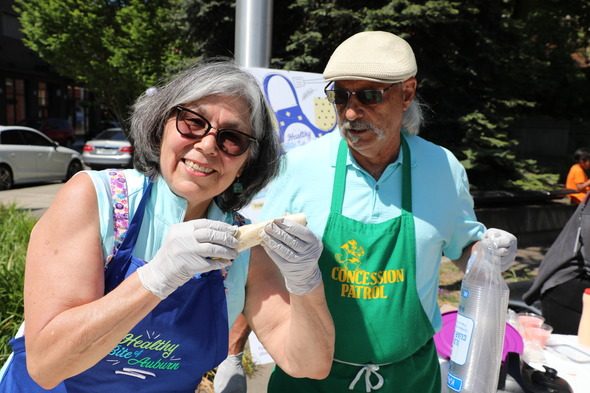 Woman in sunglasses and apron smiling while holding food, standing beside a man in a green apron at an outdoor event.