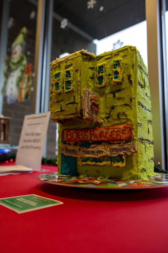 Gingerbread house modeled after Bob’s Burgers, decorated in bright green icing and displayed on a red table.