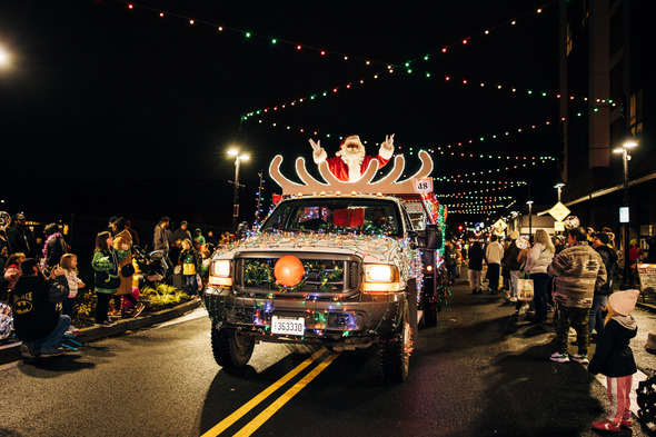 Santa rides atop a festively lit vehicle during a nighttime holiday parade as families line the street.