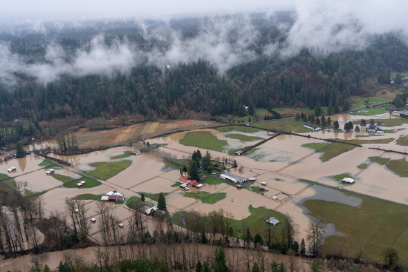 Floodwaters cover farmland, barns, and fields near a wooded hillside.