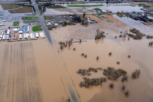 Aerial view of flooded farmland and a roadway submerged by muddy floodwaters near buildings and trees.