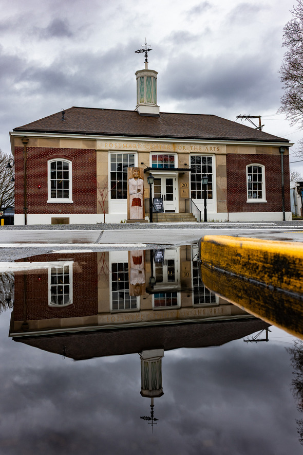 Historic brick Postmark Center for the Arts building reflected in a puddle on a cloudy day.