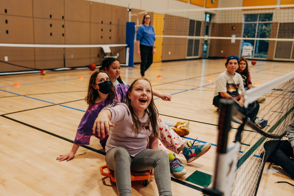 A group of kids sitting on little mobile scooter things in a gym waiting to hit a yellow balloon 