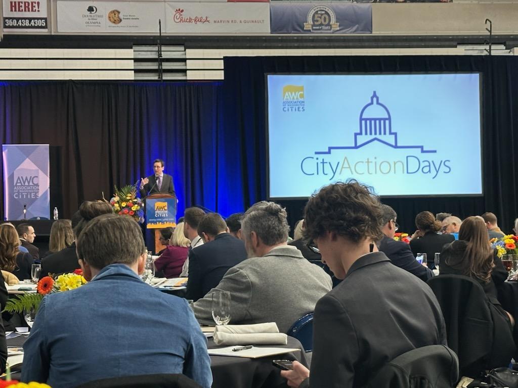 Audience seated at tables listens to a speaker on stage during AWC City Action Days, with a large screen displaying the event logo.