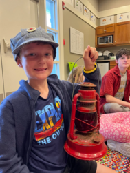 A kid smiling wearing a hat and holding one of those old fashioned lanterns 