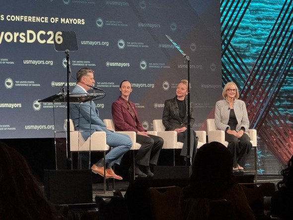 Panelists seated on stage at the U.S. Conference of Mayors event with microphones and teleprompters.