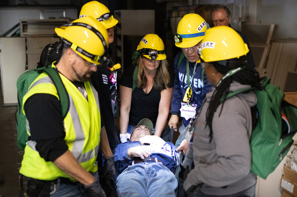 CERT volunteers in yellow helmets practice emergency rescue drill carrying a mock patient on a stretcher.