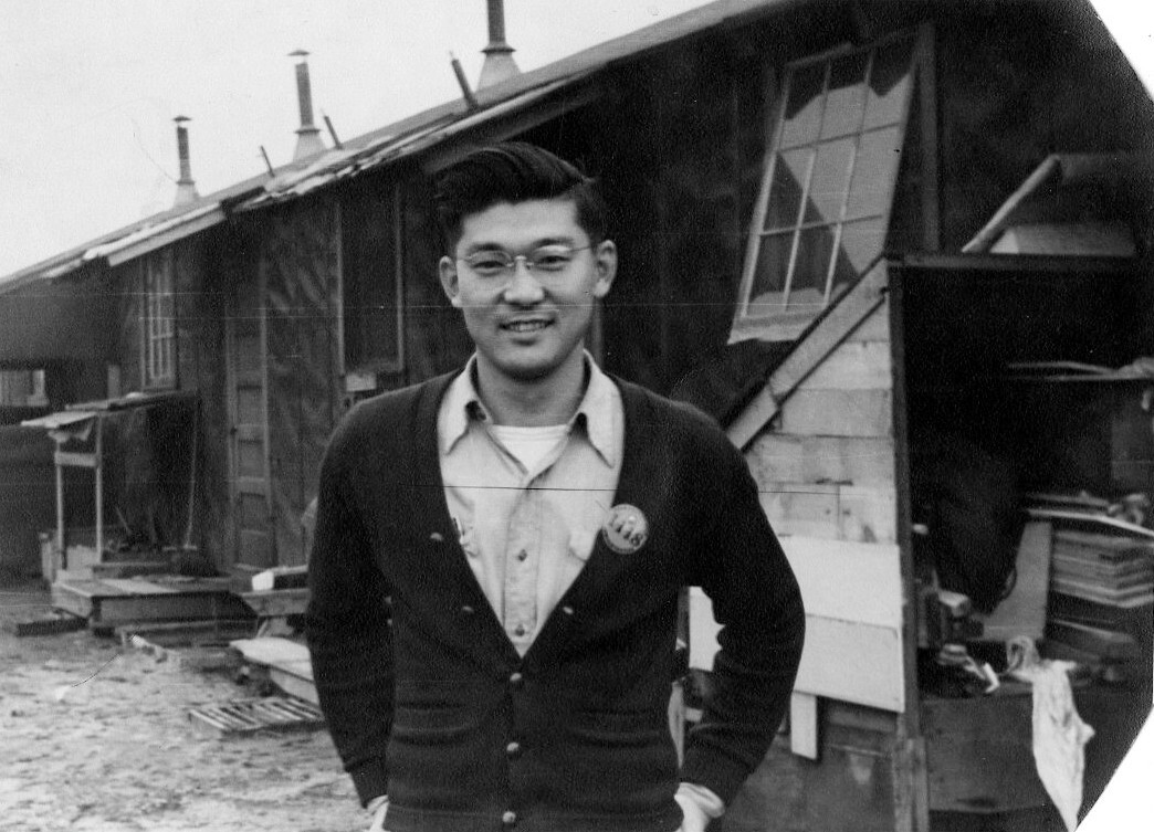Historic black-and-white portrait of a young man standing outside a row of barracks-style buildings.