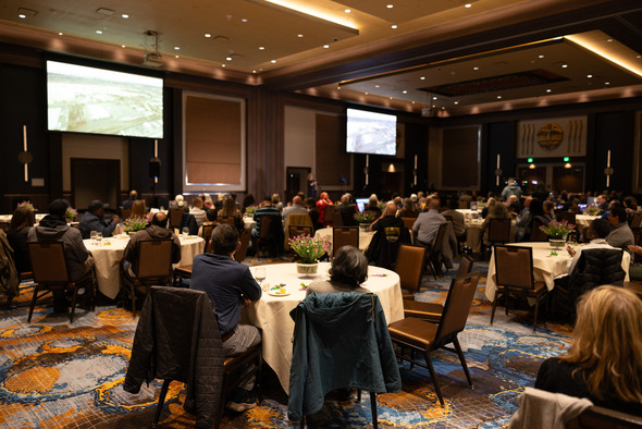 Audience seated at round tables during State of the City address, watching presentation screens in ballroom.