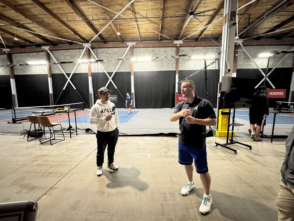 two men standing in a facility like area with some pickelball courts talking 